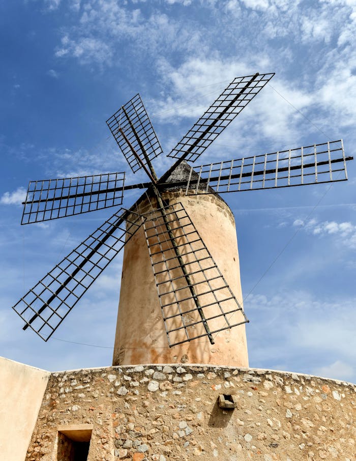 A stone windmill with metallic arms stands tall under a bright blue sky in rural Spain.